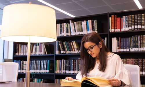 A young girl with glasses sitting at the library table reading a book under the lamp light