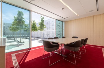 A meeting room with limited seating in Aga Khan Centre, tables and chairs placed on a red carpet facing the wall on one side and an AKC garden view on the other .